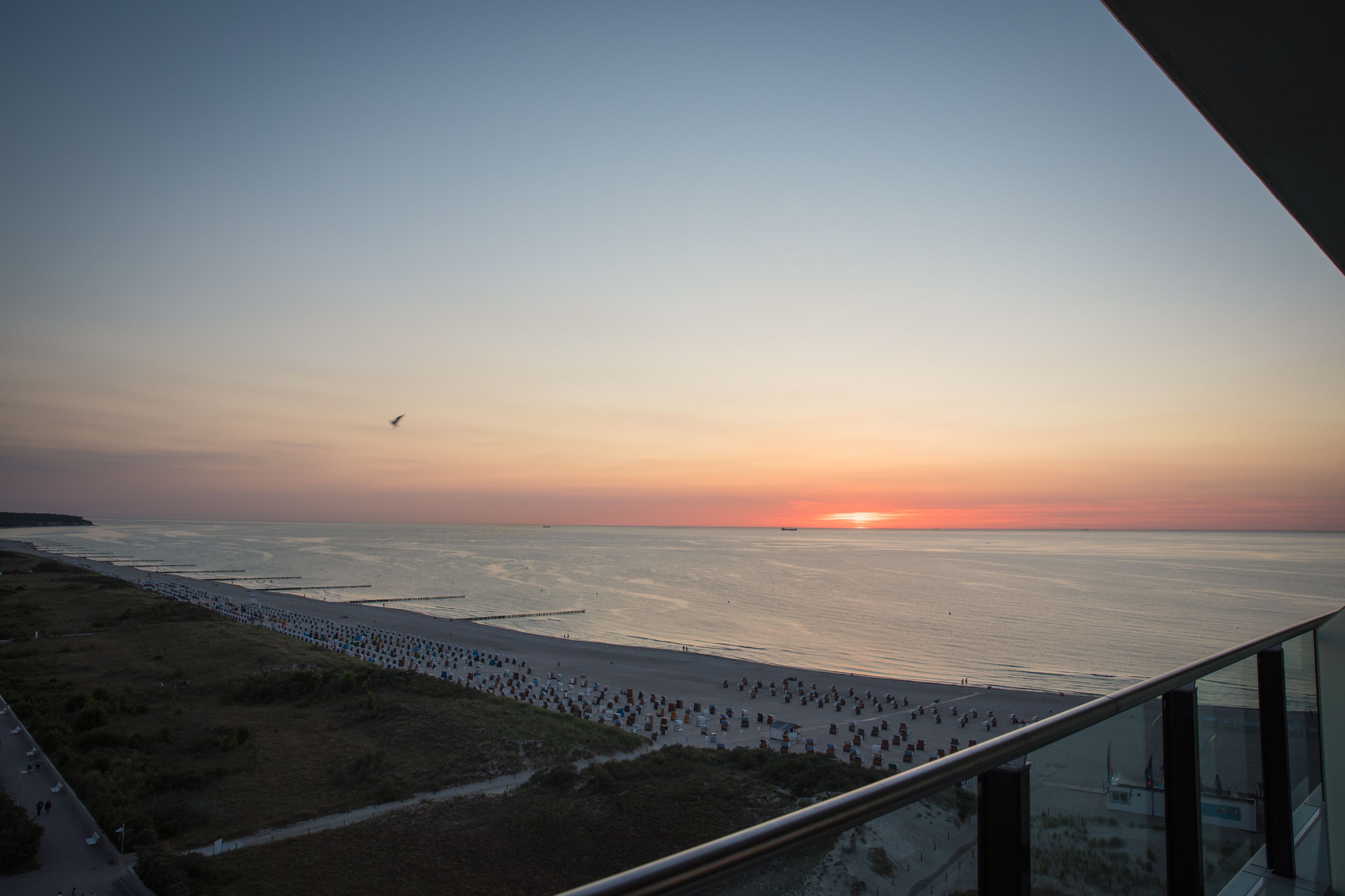 Vista del atardecer desde un balcón con vista a una playa donde se ven personas caminando y un muelle que se adentra en el agua.