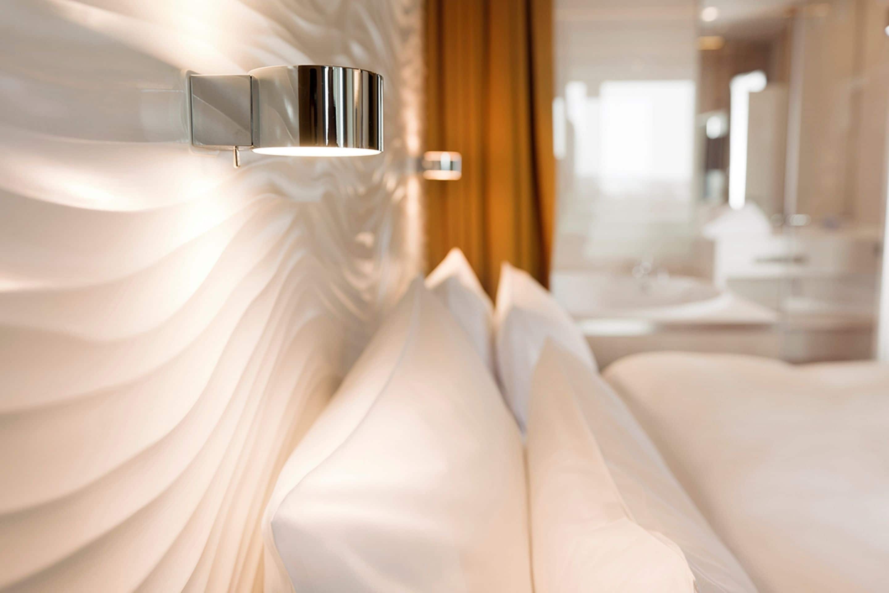 Une chambre d’hôtel moderne, dotée d’un élégant luminaire mural métallique, d’un lit blanc moelleux agrémenté de plusieurs oreillers, et où l’on aperçoit une salle de bain à l’arrière-plan.