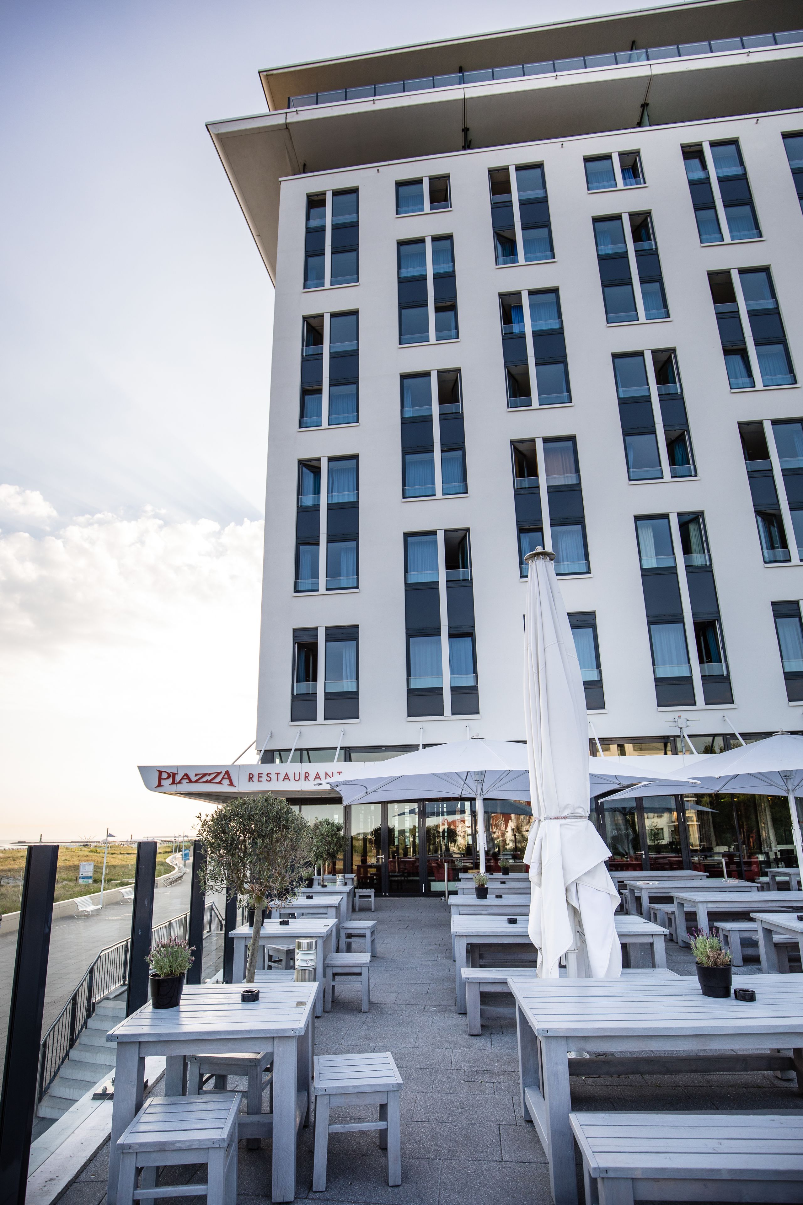 Un edificio moderno con un restaurante llamado "Piazza Restaurant" en la planta baja. Hay mesas y sillas dispuestas al aire libre para comer.