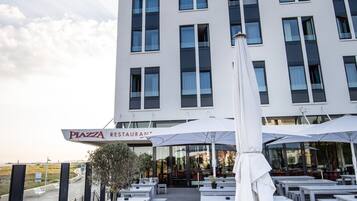 Il s’agit d’un bâtiment moderne qui abrite au rez-de-chaussée un restaurant appelé « Piazza Restaurant ». Des tables et des chaises sont disposées à l’extérieur pour permettre aux clients de prendre leurs repas.