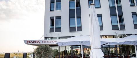 Un edificio moderno con un restaurante llamado "Piazza Restaurant" en la planta baja. Hay mesas y sillas dispuestas al aire libre para comer.