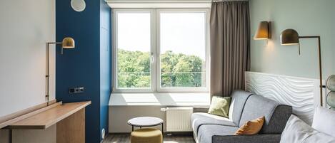 A modern hotel room with a bed, a sofa, a small table, and a desk. There are two windows with curtains, and a view of greenery outside.