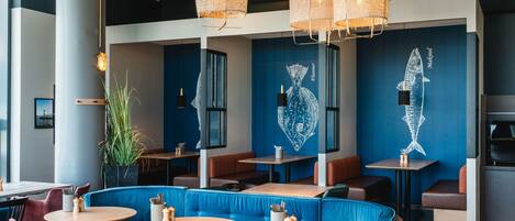 Interior de un restaurante con asientos tipo banca en azul, mesas de madera y lámparas de mimbre colgantes.