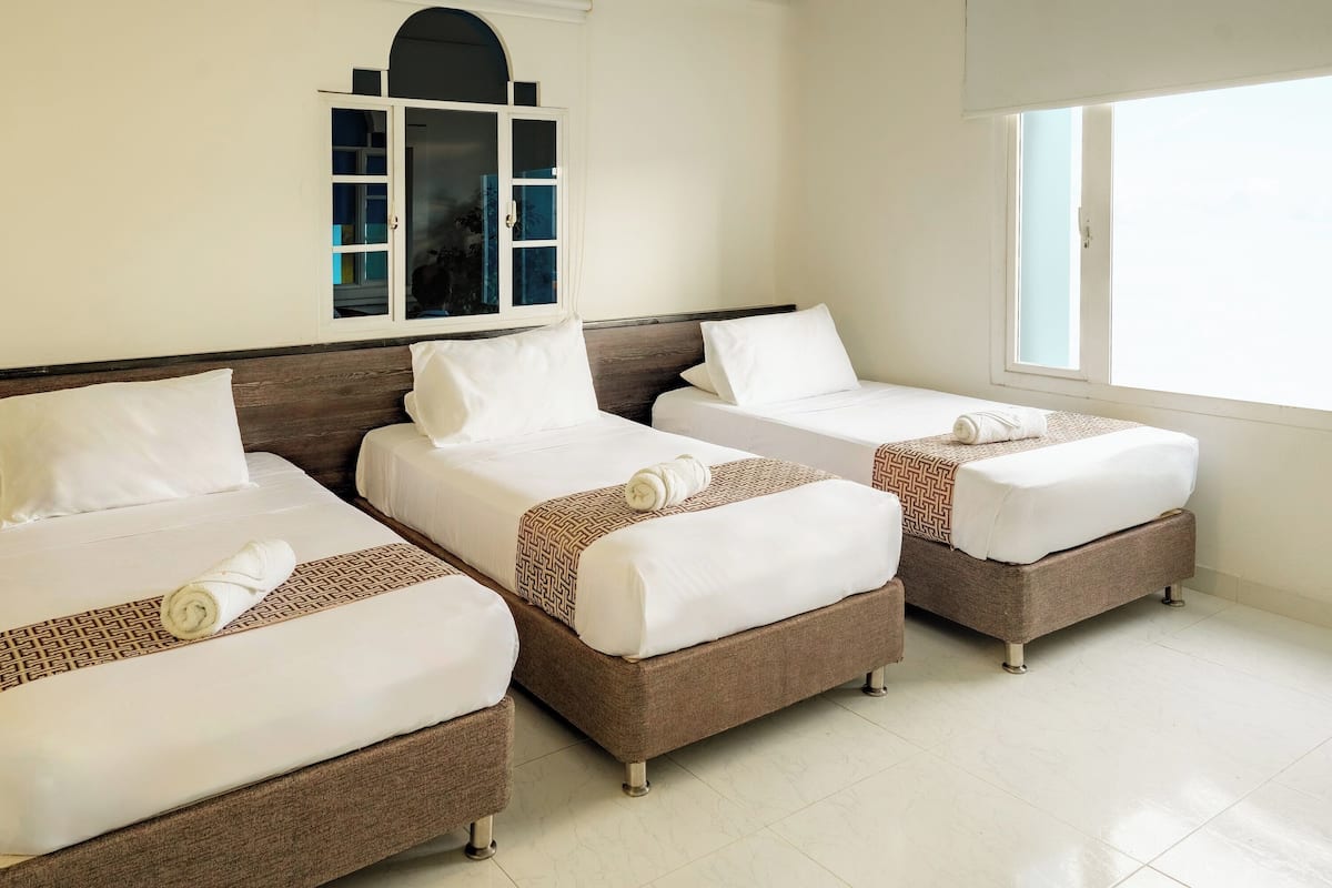 A hotel room with three single beds, each with white linens and a rolled towel. The room has a large window on the right and a built-in cabinet with glass doors on the left.
