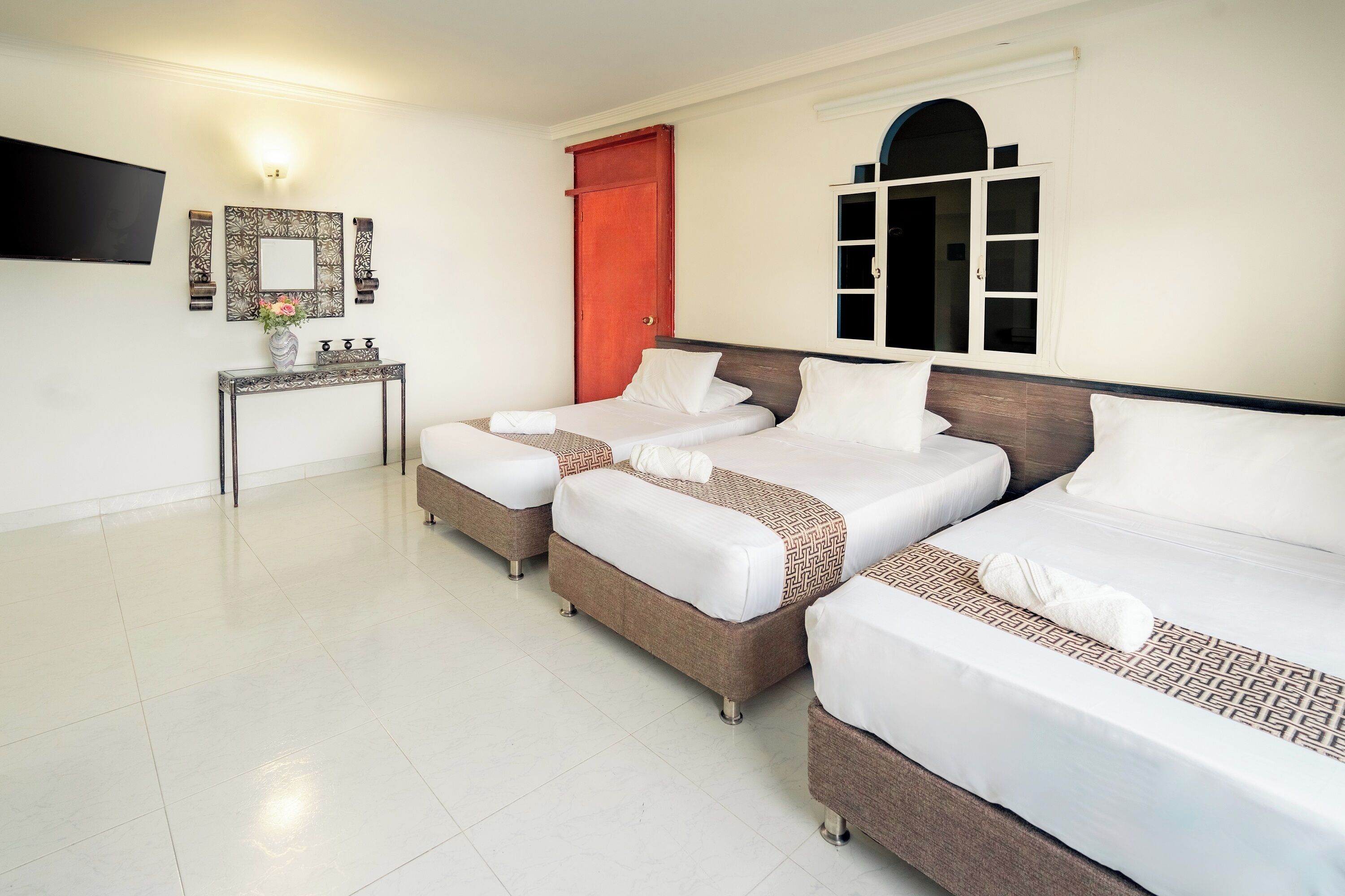 Habitación de hotel con tres camas, un televisor, una consola con un florero y una puerta roja.