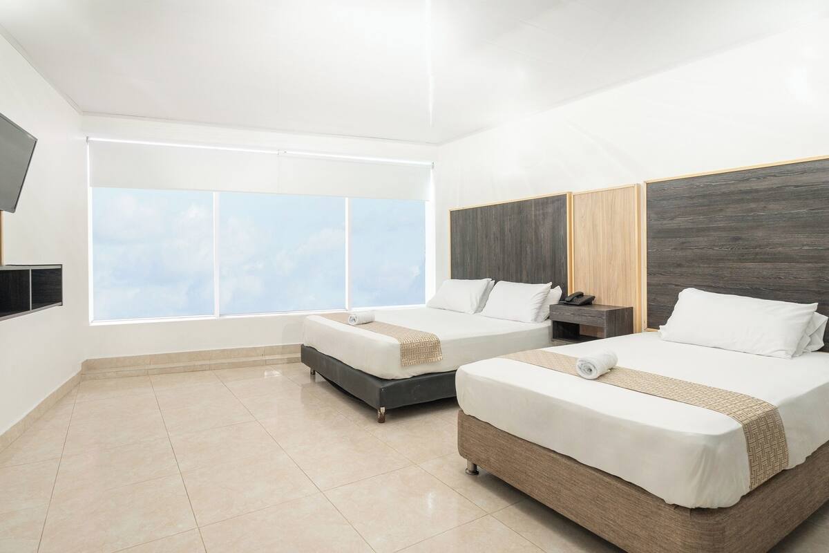 A modern hotel room with two beds, a TV mounted on the wall, and a large window offering a view of the sky.