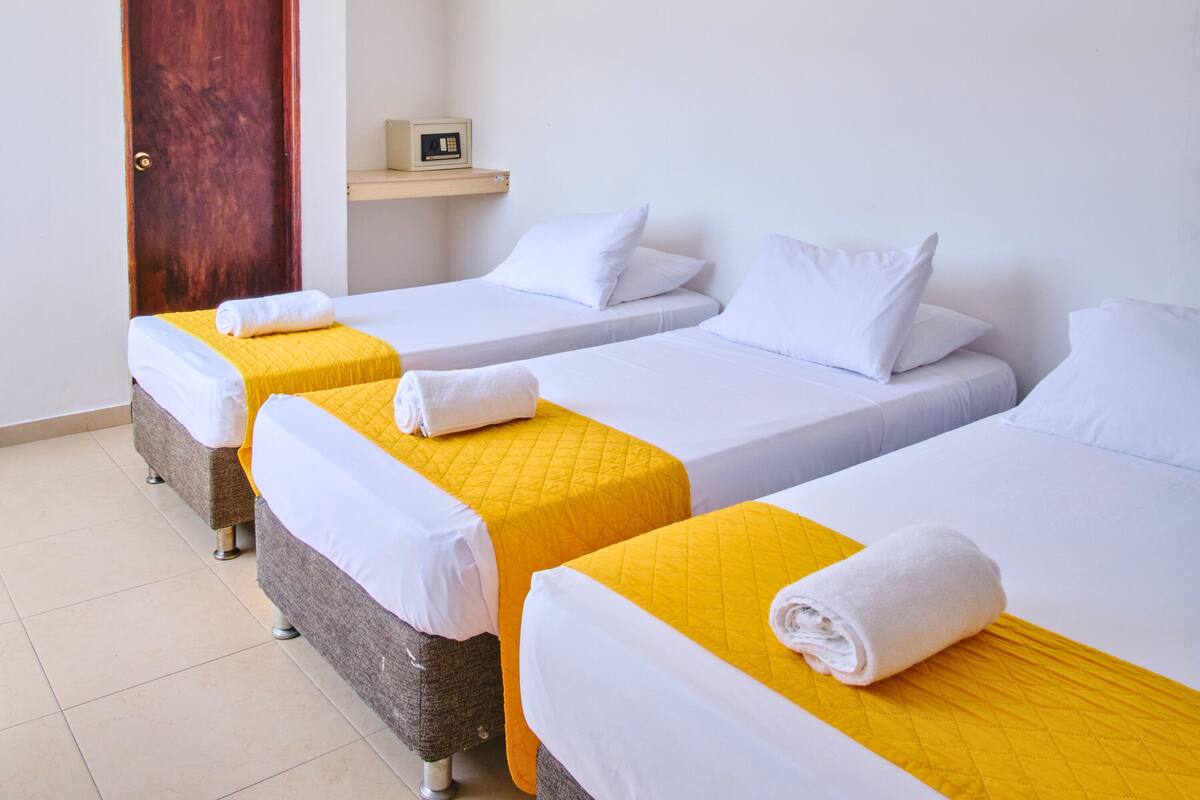 A hotel room with three single beds, each with a white and yellow bedspread and a rolled white towel. There is a wooden door and a small shelf with a clock.
