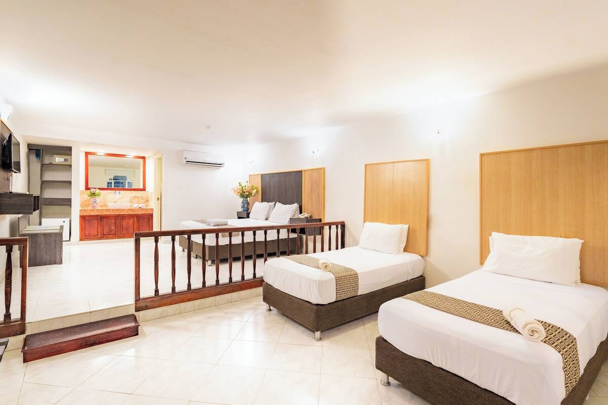 A hotel room with two beds, a wooden headboard, a small desk with a chair, a balcony with a railing, and a bathroom with a mirror and sink.