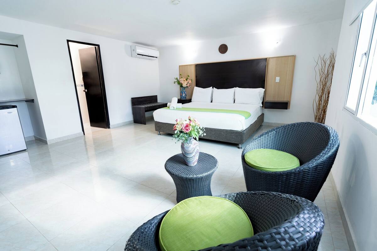 A modern hotel room with a large bed, a small table with a vase of flowers, wicker chairs, and a view of the outside.