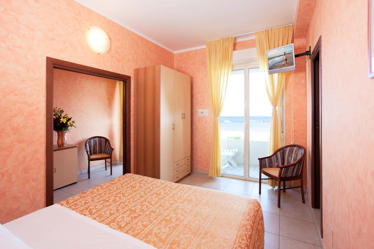 A hotel room with a bed, wardrobe, two chairs, a small table with a vase, and a view of the sea through a sliding door.
