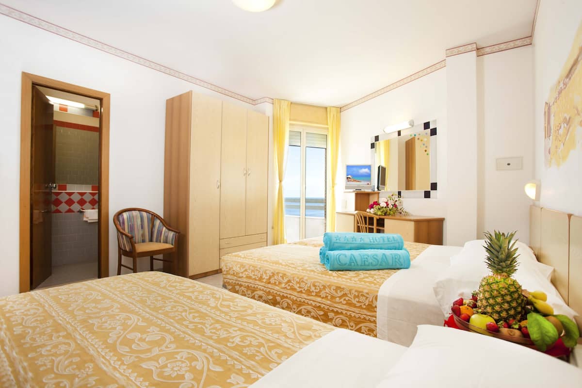A hotel room with two beds, a chair, a desk with a computer, a mirror, and a view of the sea.