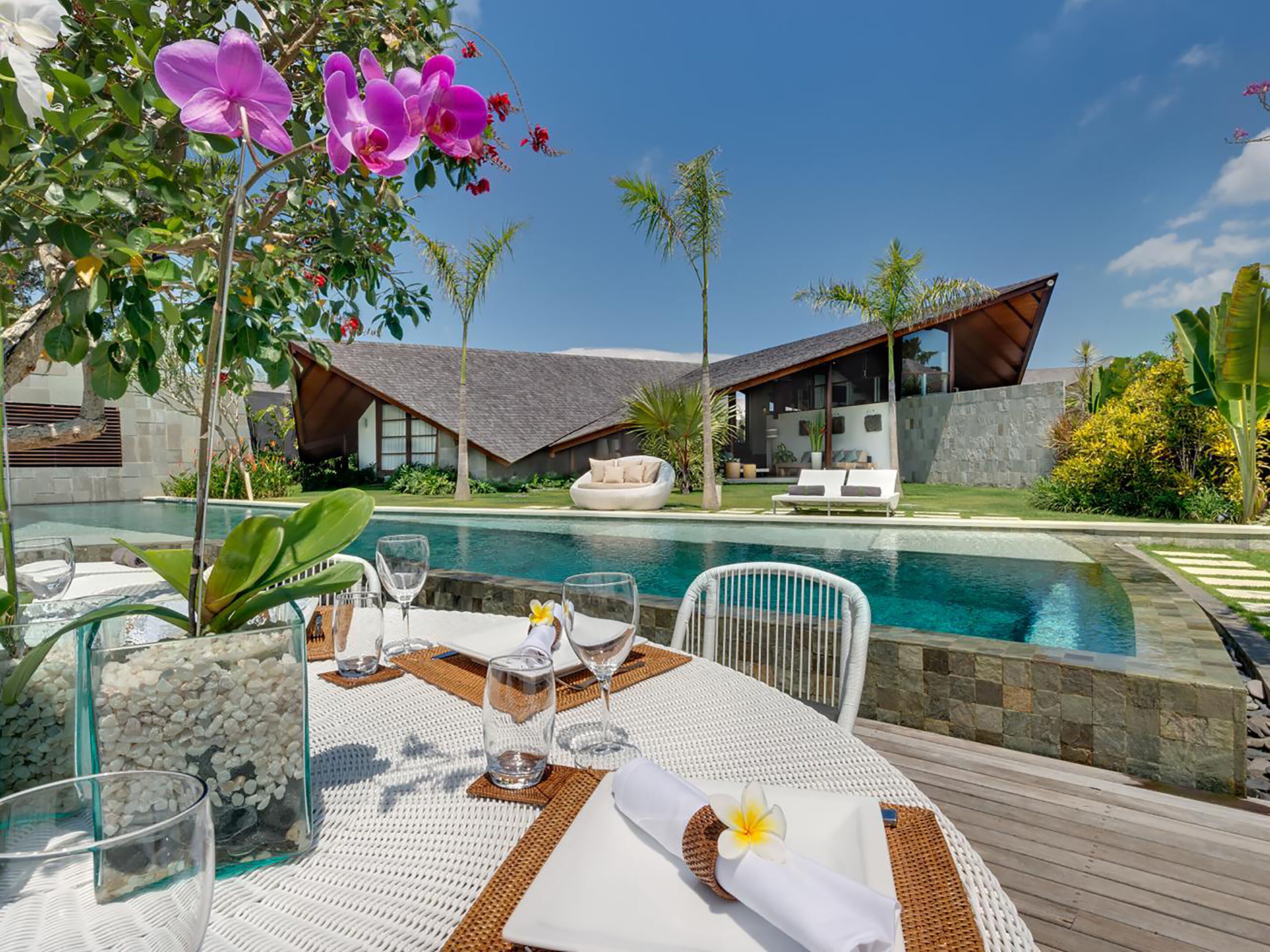 A poolside dining area with a table set for two, surrounded by a pool and a modern house in the background.