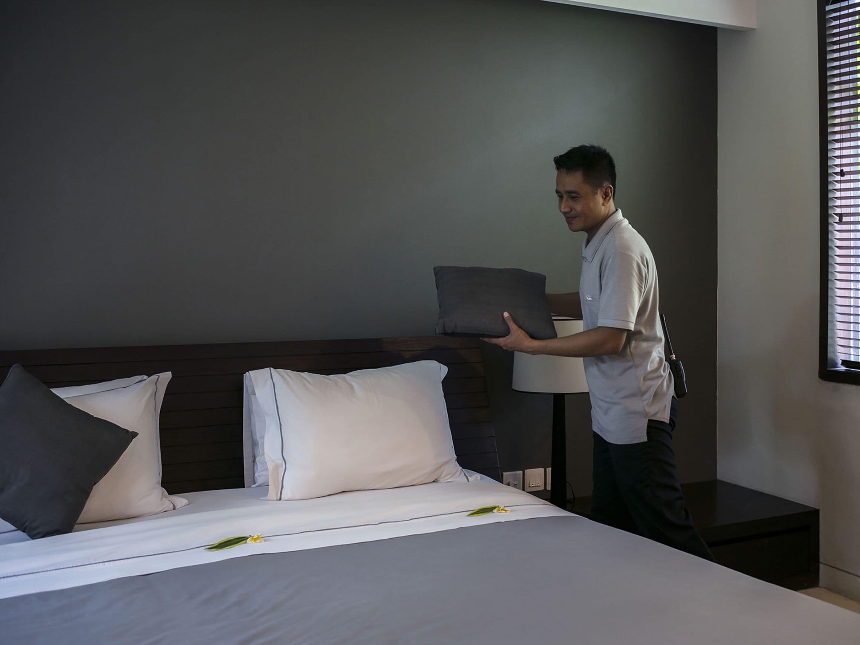 A hotel room with a neatly made bed, a bedside lamp, and a window with blinds.