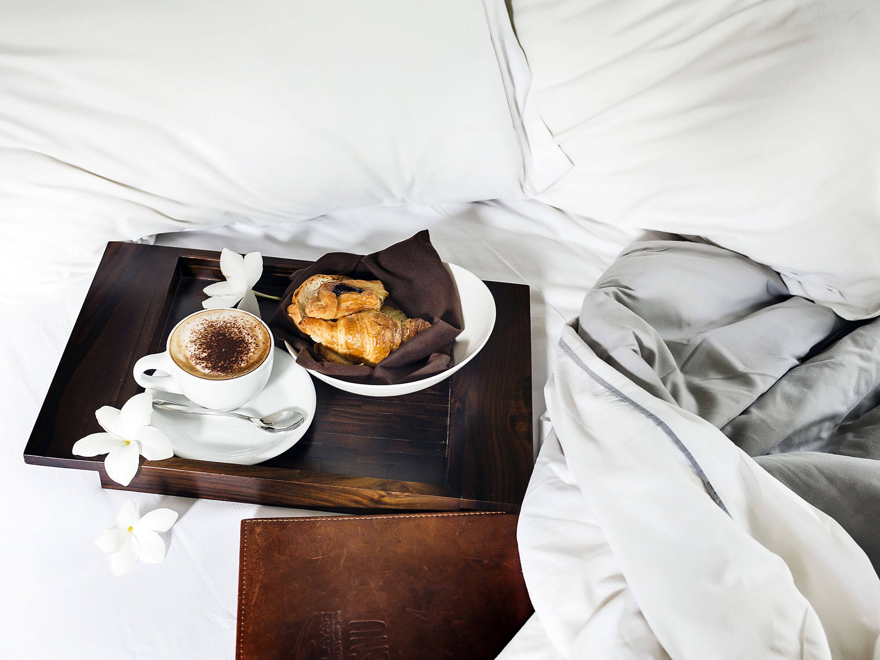 Uma bandeja de café da manhã bem organizada com uma xícara de café, croissants e uma tigela de doces, sobre uma bandeja de madeira, colocada sobre uma cama com roupa de cama branca.