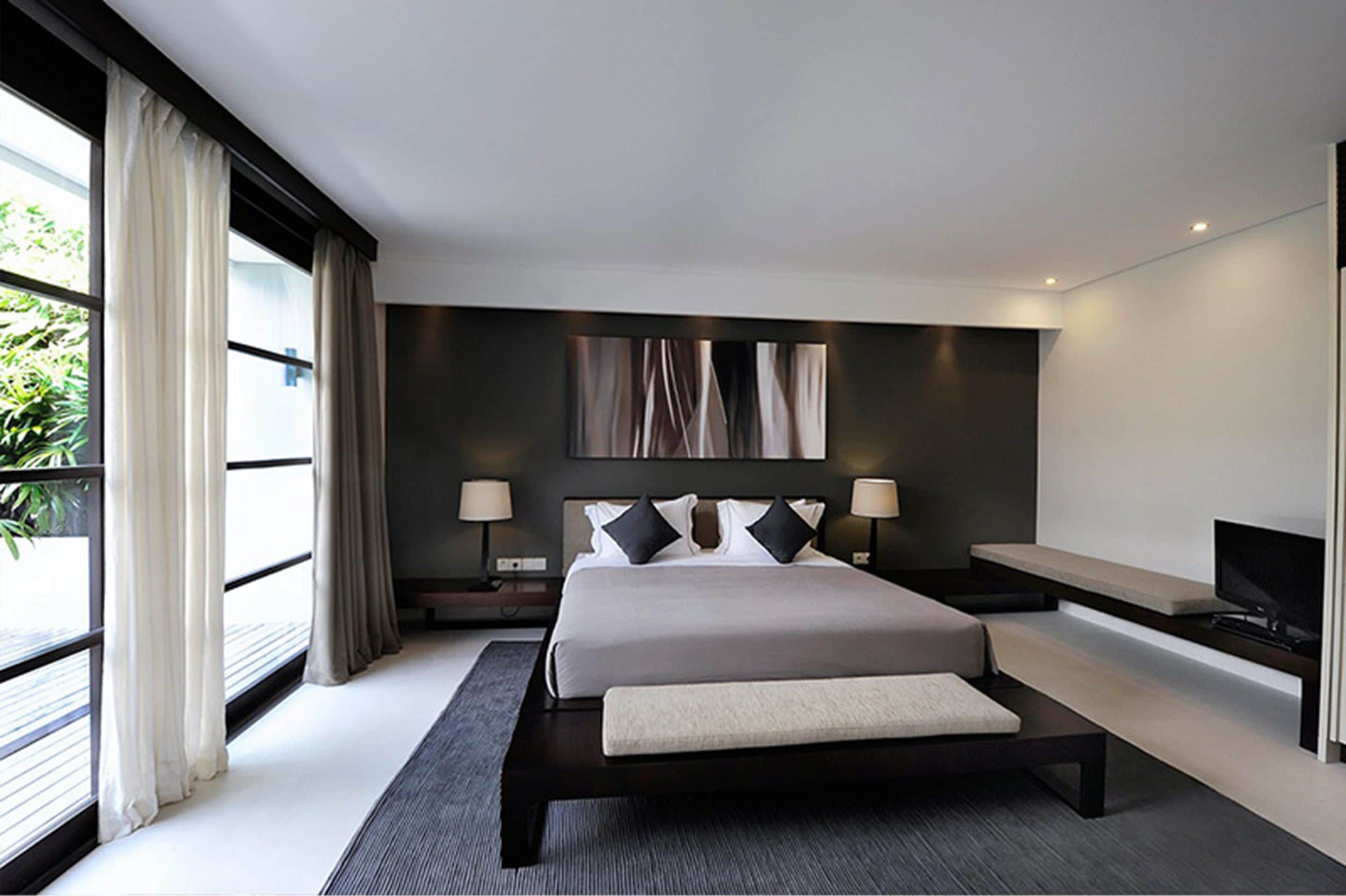 A modern bedroom with a large bed, bedside lamps, a TV, and a view of greenery outside.