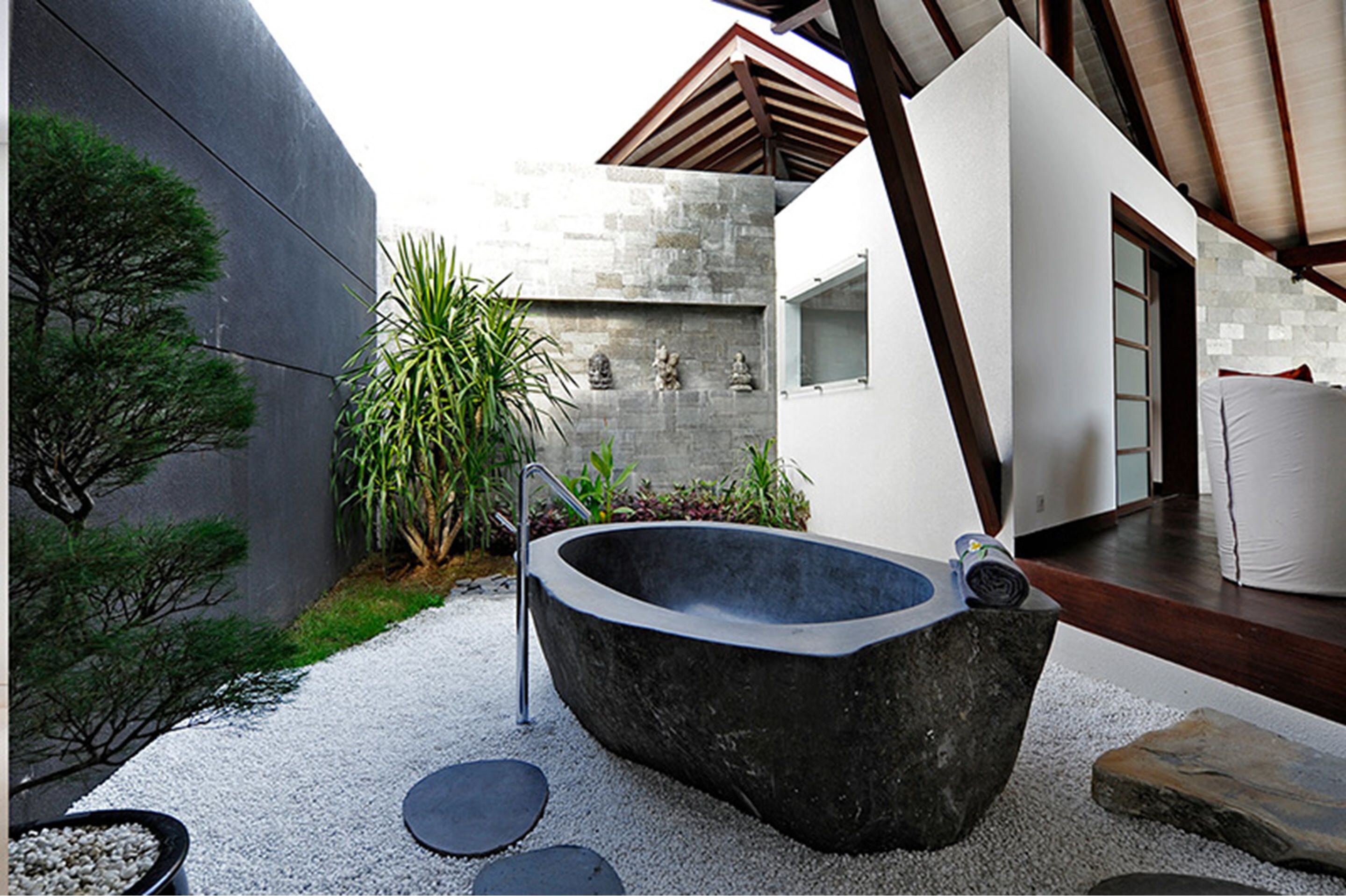 A modern outdoor spa area with a large stone hot tub, surrounded by gravel, plants, and a wooden deck.