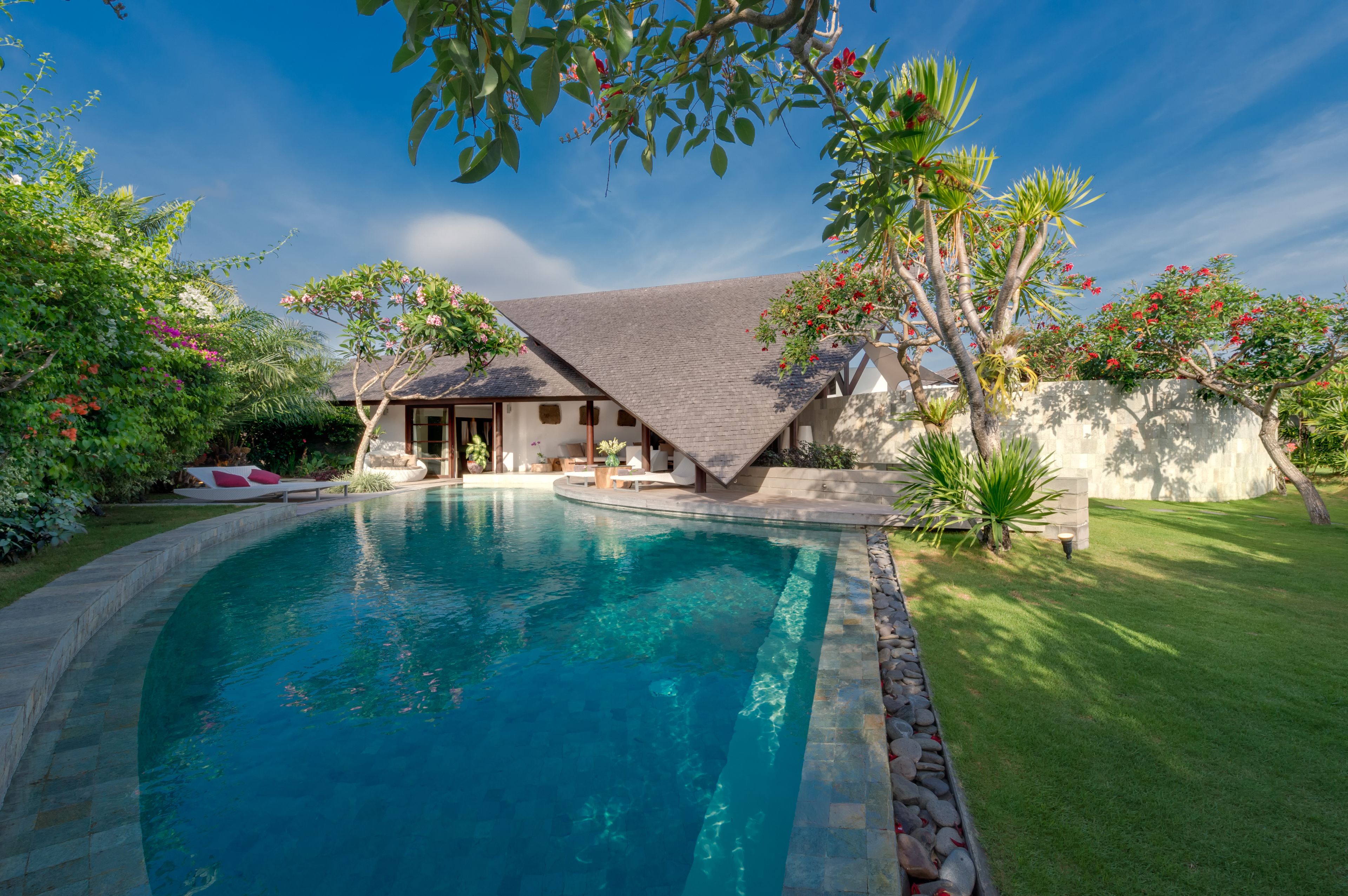 A swimming pool with a house in the background, surrounded by lush greenery and a clear blue sky.