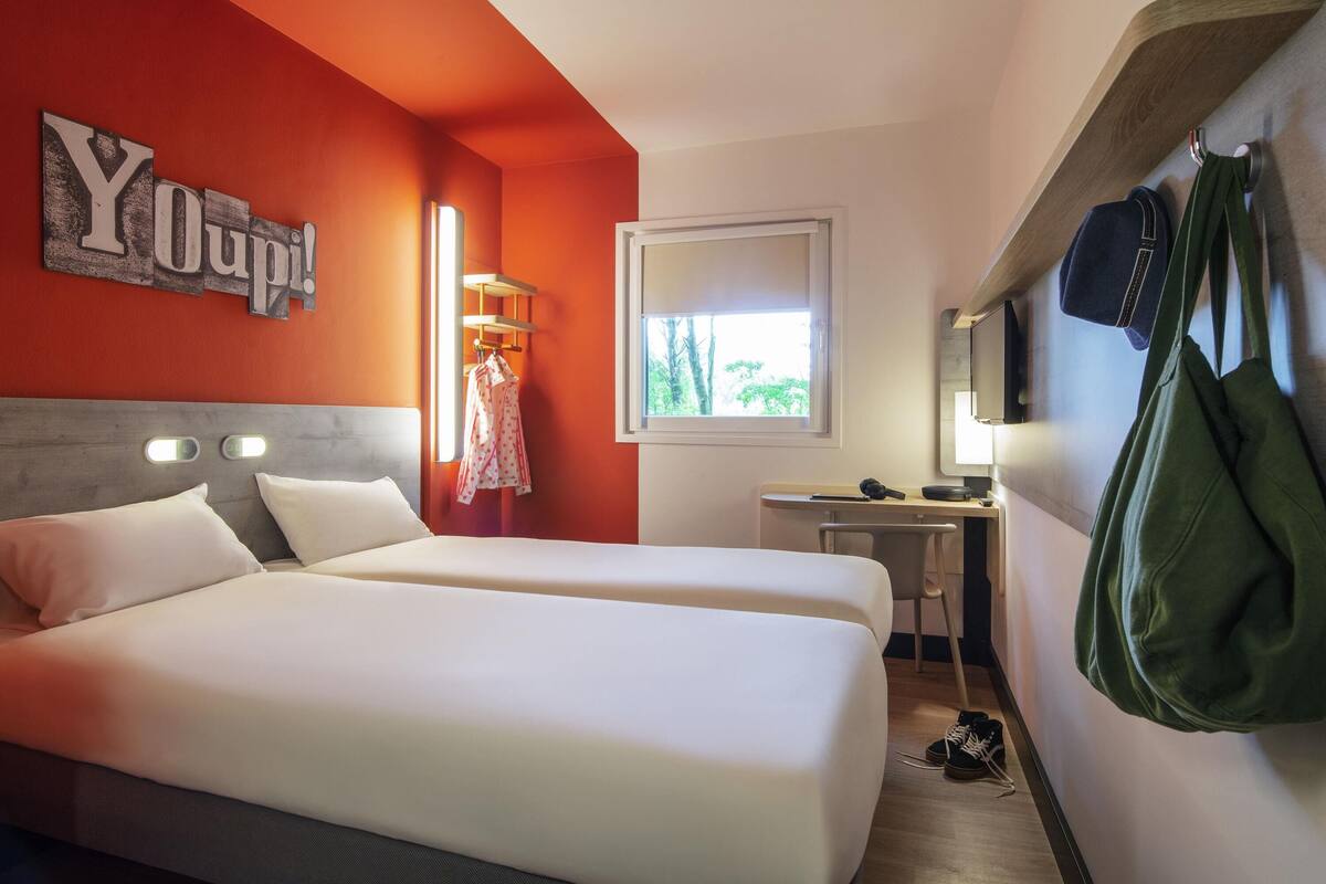 Ein Hotelzimmer mit zwei Betten, einem Schreibtisch, einem Stuhl, einem Fenster mit Blick auf Bäume und einem an der Wand befestigten Kleiderhaken mit Taschen.