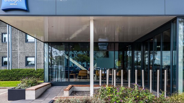 Un bâtiment moderne avec une façade en verre, une enseigne indiquant la présence d’hôtels ibis budget, et une vue dégagée sur la rue.