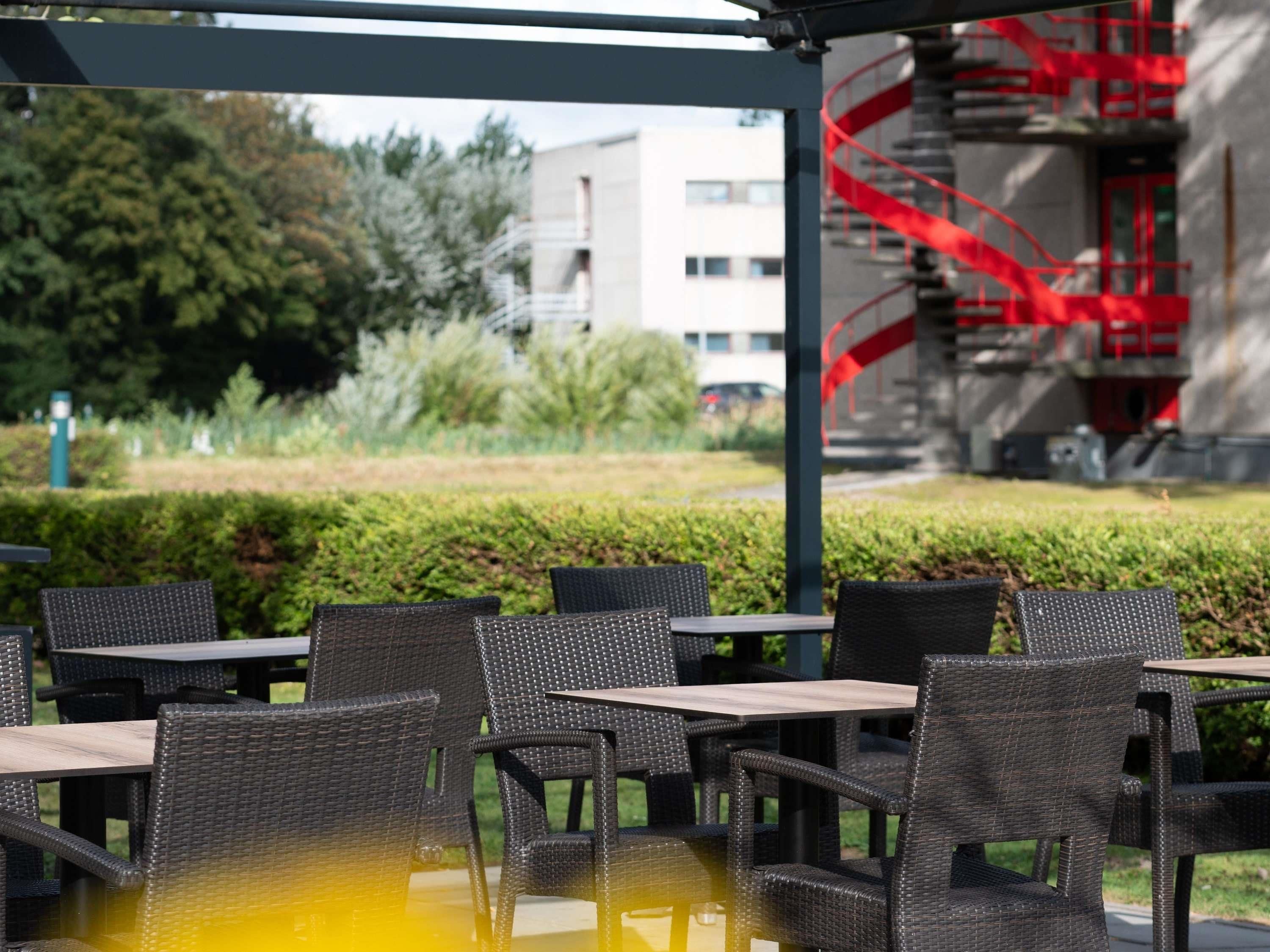 An outdoor seating area with wicker chairs and tables, a glass-top table, and a view of a building with a red spiral staircase.
