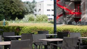 Een terras met rieten stoelen en tafels, een tafel met glazen blad en uitzicht op een gebouw met een rode spiraaltrap.