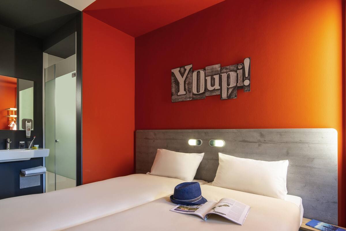 Ein Hotelzimmer mit einem Bett, einem Nachttisch, einem Spiegel, einer roten Wand und einem Schild mit der Aufschrift 'Youpi!'.