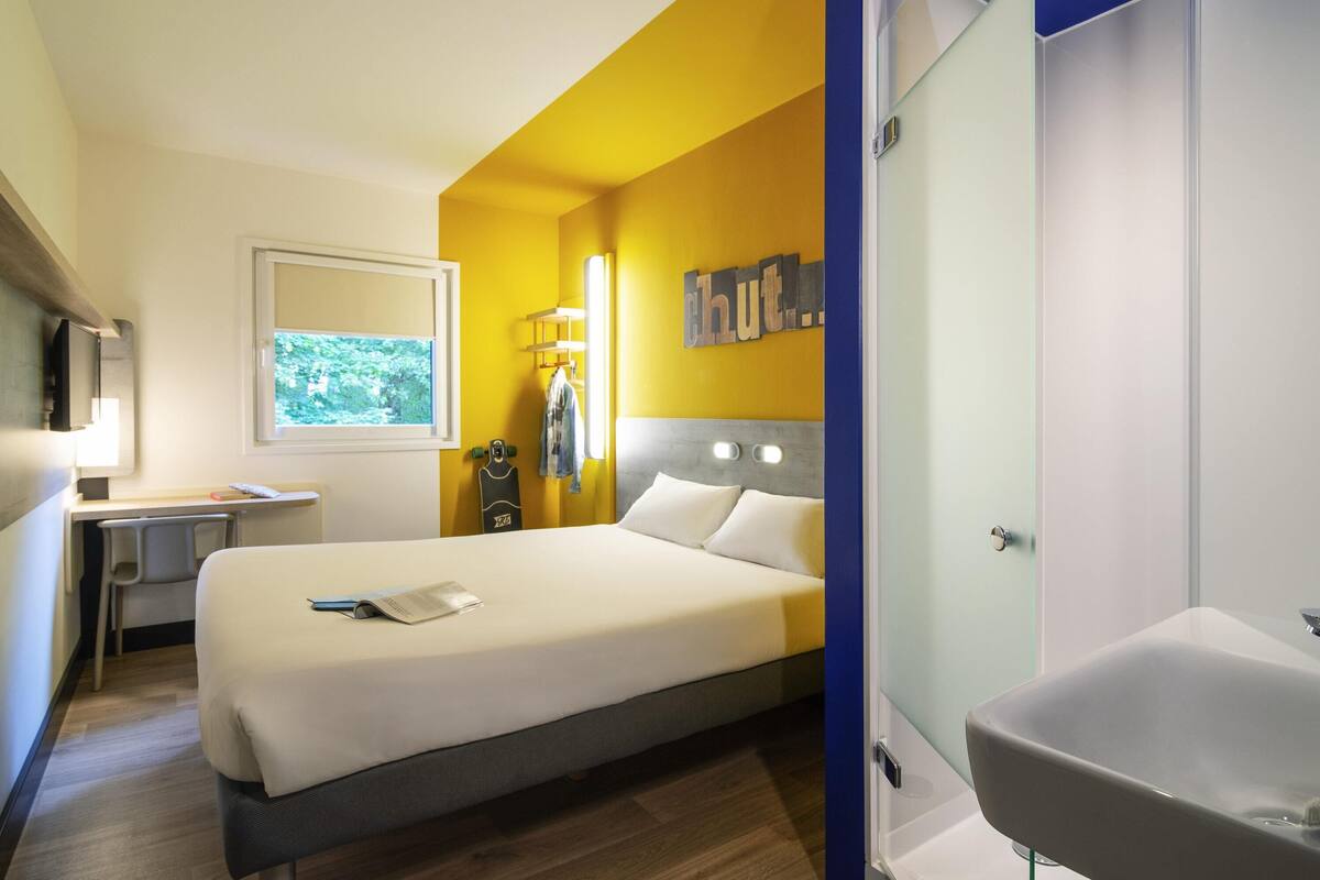 Ein Hotelzimmer mit einem Bett, einem Schreibtisch, einem Stuhl, einem Fenster, einem an der Wand befestigten Fernseher und einem Badezimmer mit Waschbecken und Spiegel.