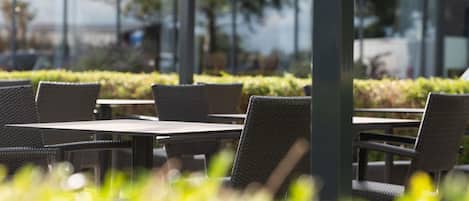 An outdoor seating area with tables and chairs, surrounded by greenery and a building in the background.