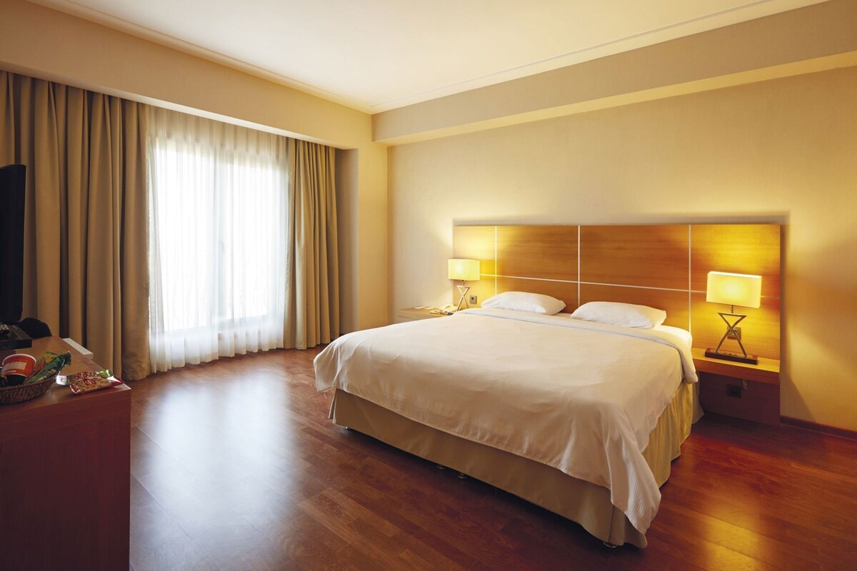 A hotel room with a large bed, two bedside tables with lamps, a wooden headboard, and a large window with curtains.