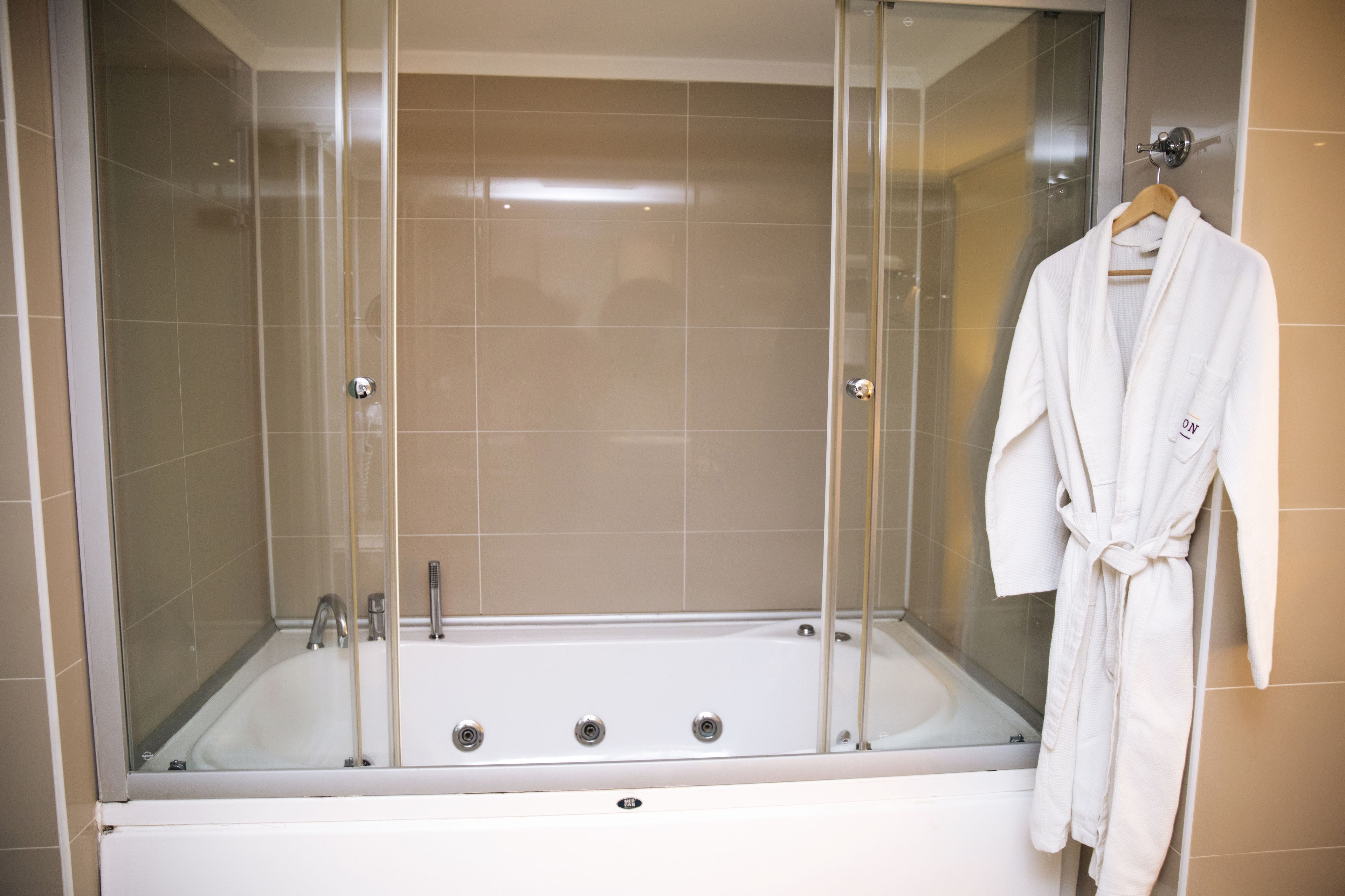 Un baño moderno con una bañera de hidromasaje con mampara de vidrio, un perchero de madera y un grifo visible.