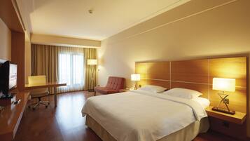 A hotel room with a large bed, a desk with a TV, a chair, and a desk lamp.