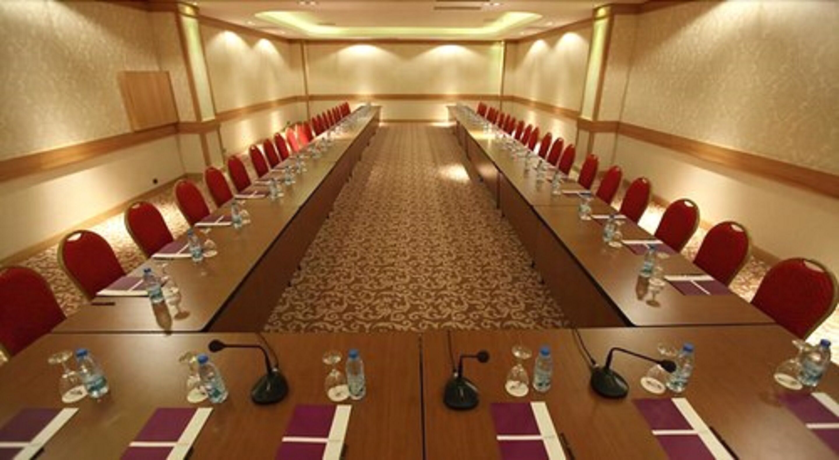 A conference room with a long table, red chairs, microphones, and water bottles.
