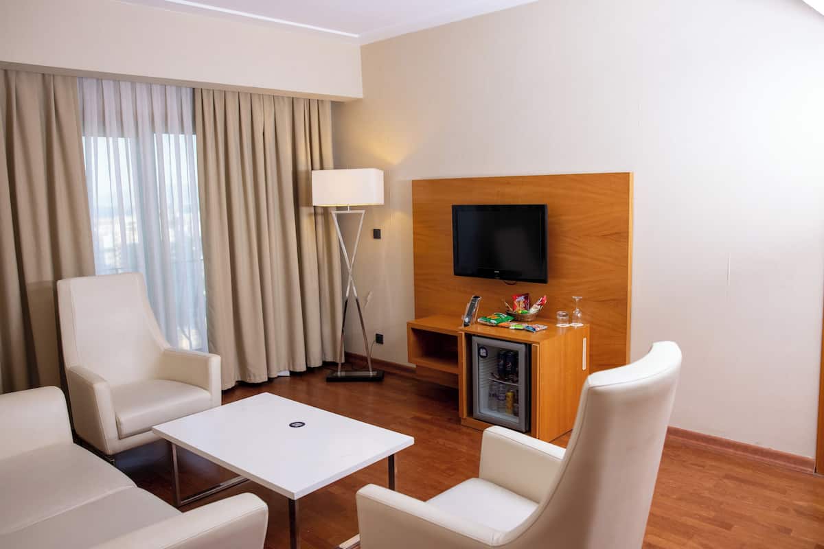 Una habitación de hotel moderna con una televisión de pantalla plana montada en un panel de madera, un mini refrigerador con aperitivos y bebidas, un sofá blanco, un sillón blanco y una pequeña mesa.