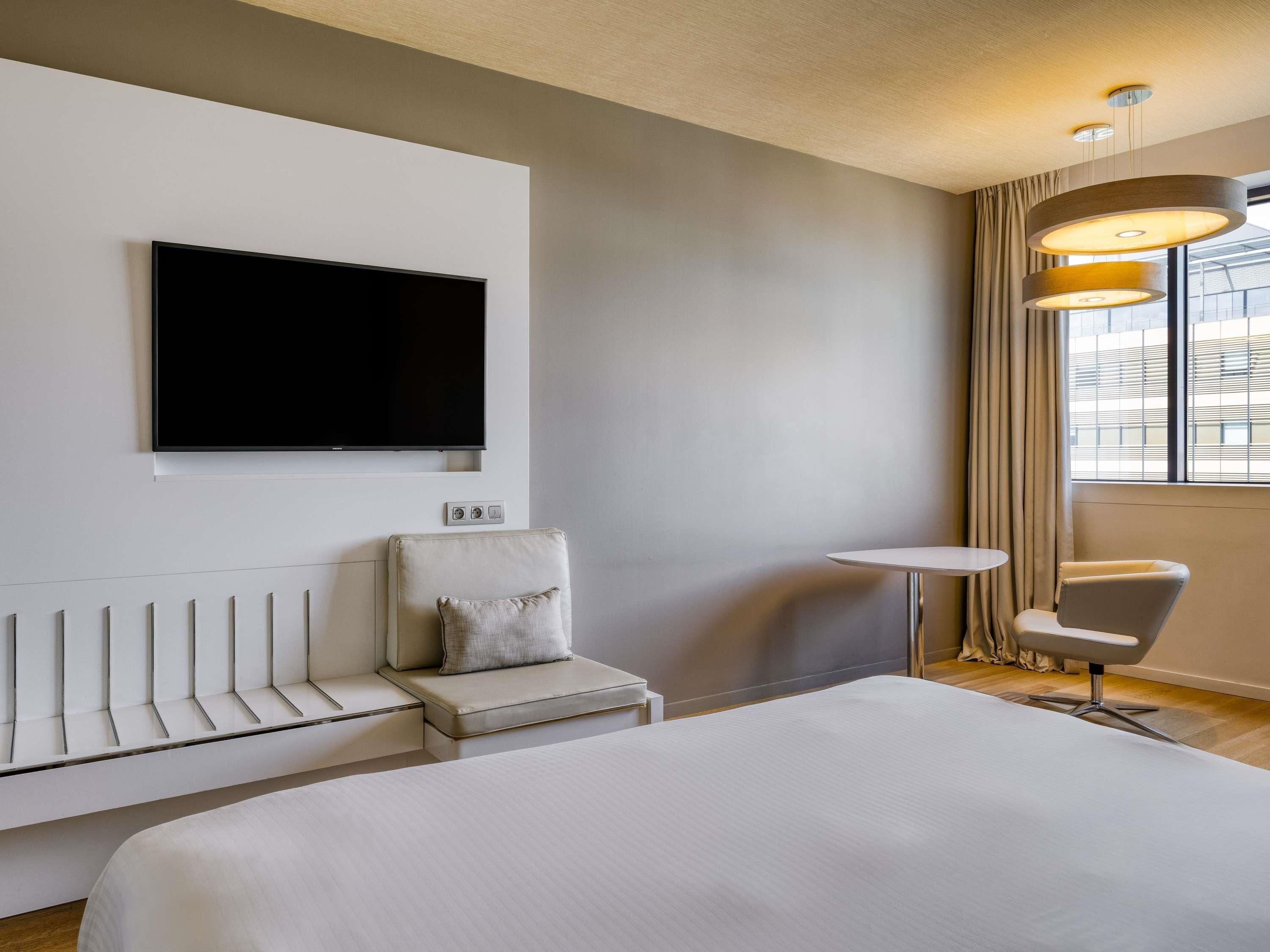 A hotel room with a bed, a TV mounted on the wall, a seating area with a chair and a small table, and a window with curtains.