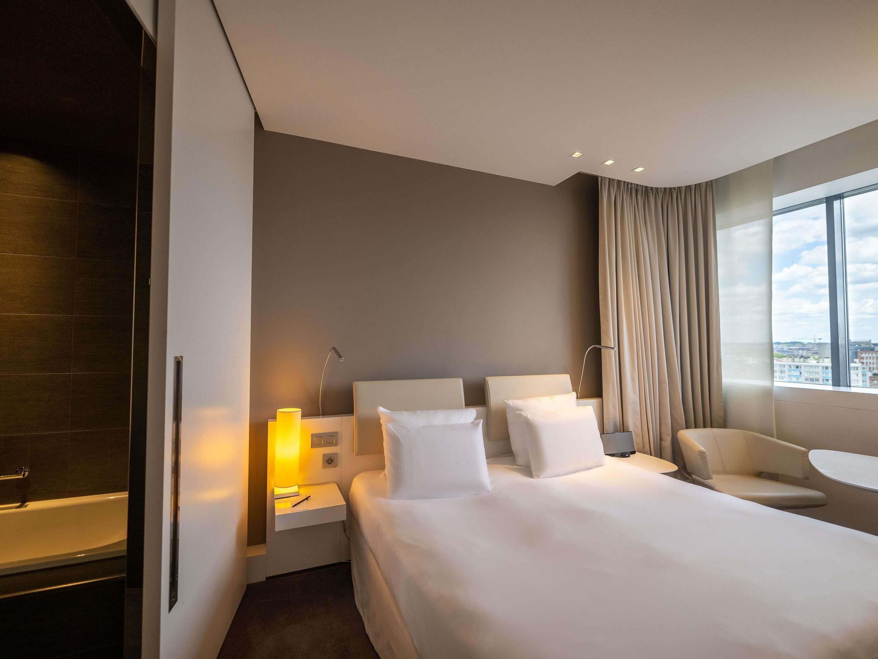 Ein modernes Hotelzimmer mit einem großen Bett, einem Schreibtisch, einem Stuhl und Blick auf die Stadt durch das Fenster.