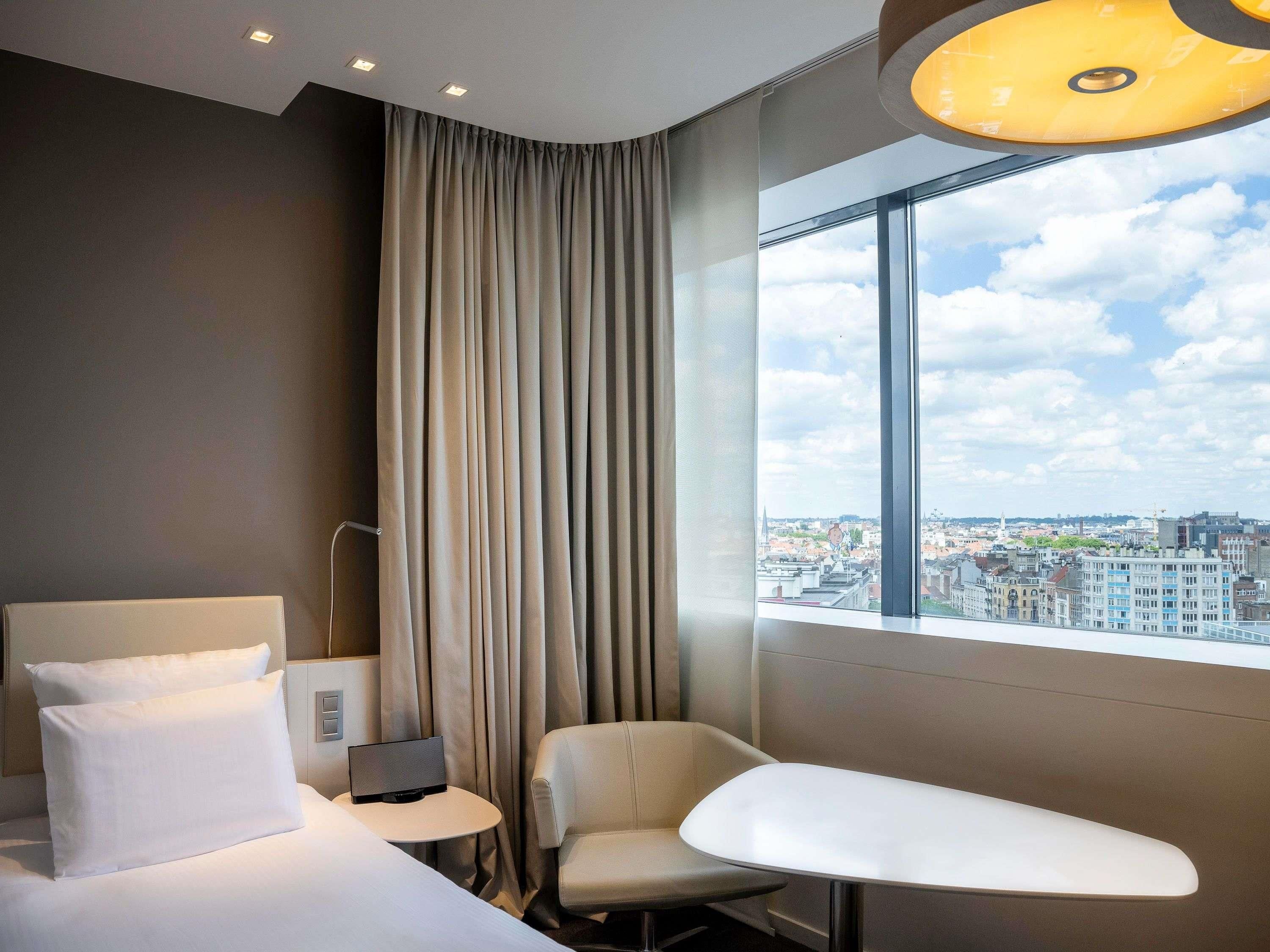 Habitación de hotel con cama, una silla, una mesita, televisor y un ventanal con vistas a la ciudad.