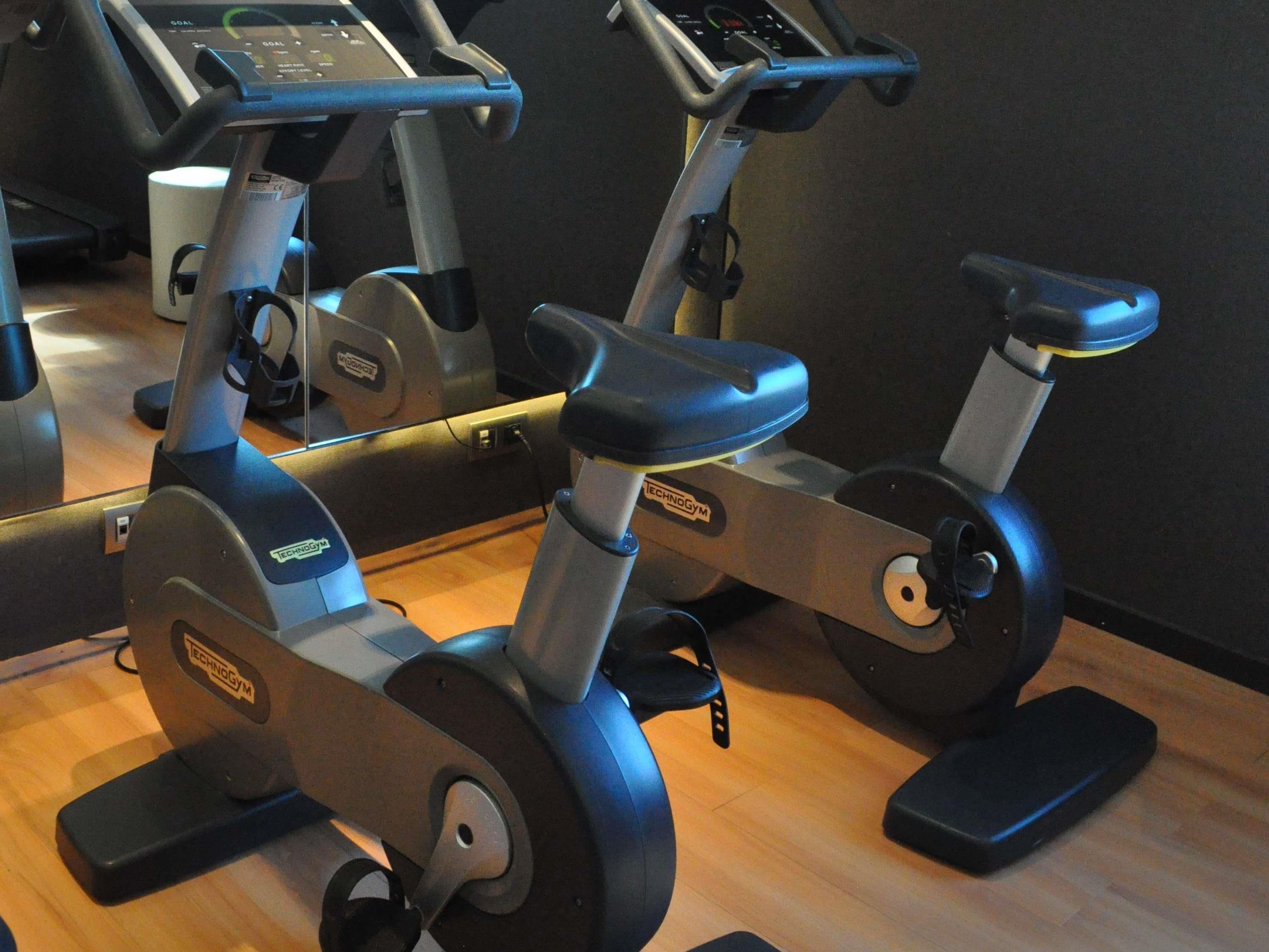 Dos bicicletas estáticas en un gimnasio, con un espejo que refleja los equipos.