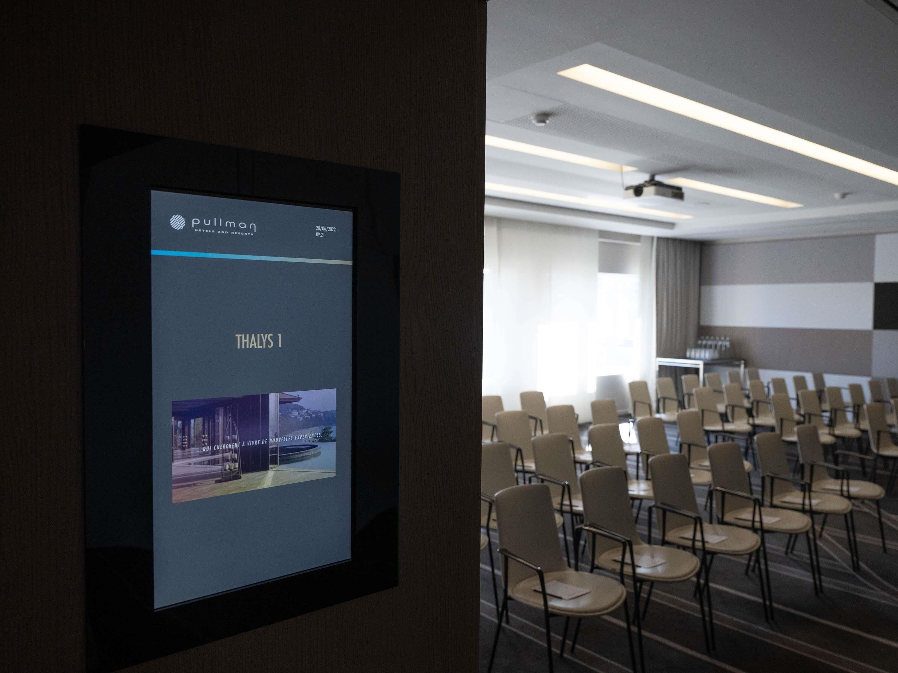 Un salón de conferencias con filas de sillas, una pantalla de proyección mostrando el nombre de un hotel y una fecha, y un diseño de techo moderno.