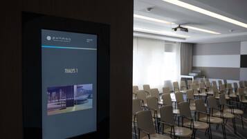 Un salón de conferencias con filas de sillas, una pantalla de proyección mostrando el nombre de un hotel y una fecha, y un diseño de techo moderno.