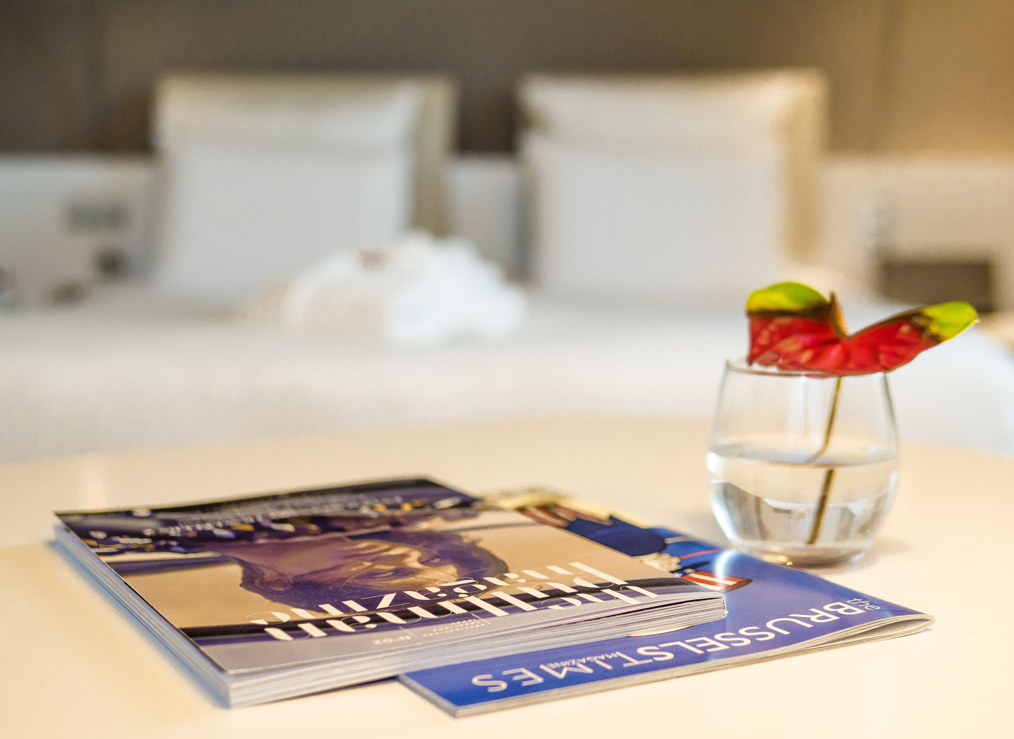 A neatly arranged hotel room with a bed, pillows, a glass of water, and a magazine on a table.