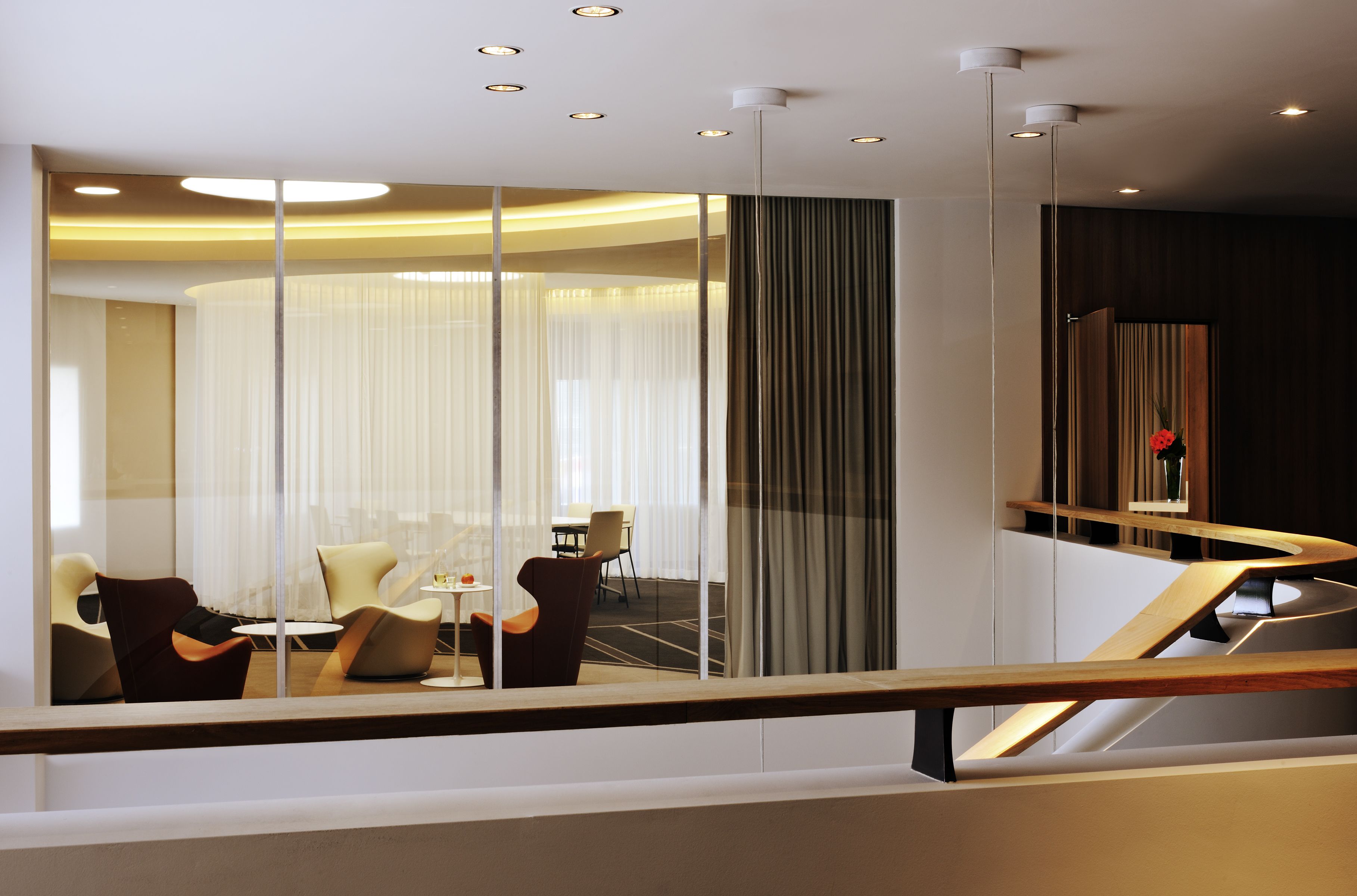 A modern hotel interior with a glass partition, a dining area with chairs and a table, and a wooden staircase.