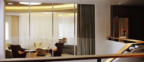 Interior de hotel moderno com divisória de vidro, área de refeições com cadeiras e mesa, e escada de madeira.