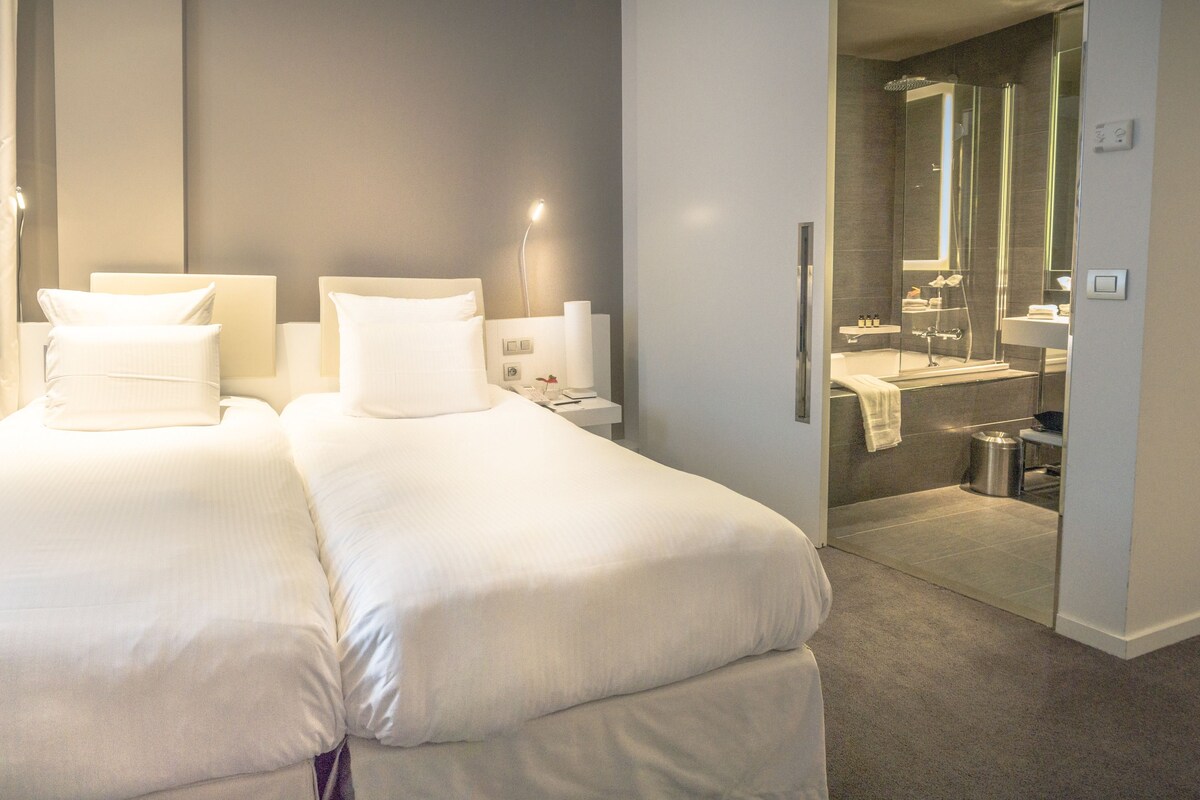 Habitación de hotel con dos camas, un escritorio, una silla y un baño visible a través de una puerta abierta.