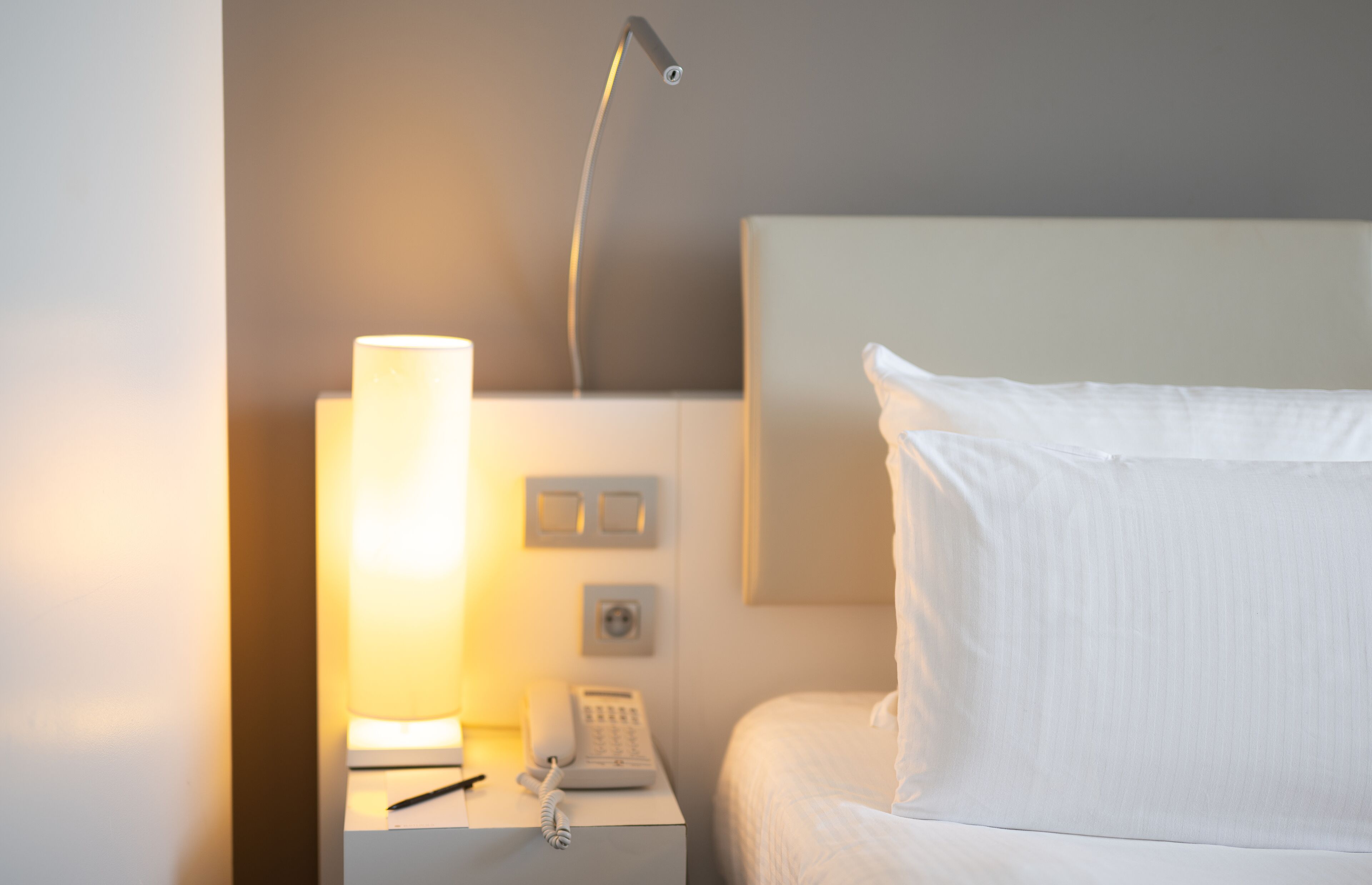 Een net opgemaakt bed met witte lakens, een bedlamp, een telefoon en een lichtschakelaar aan de muur.