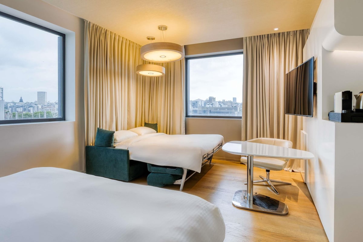 Habitación de hotel con dos camas, un escritorio, una silla, un televisor y vistas a la ciudad.