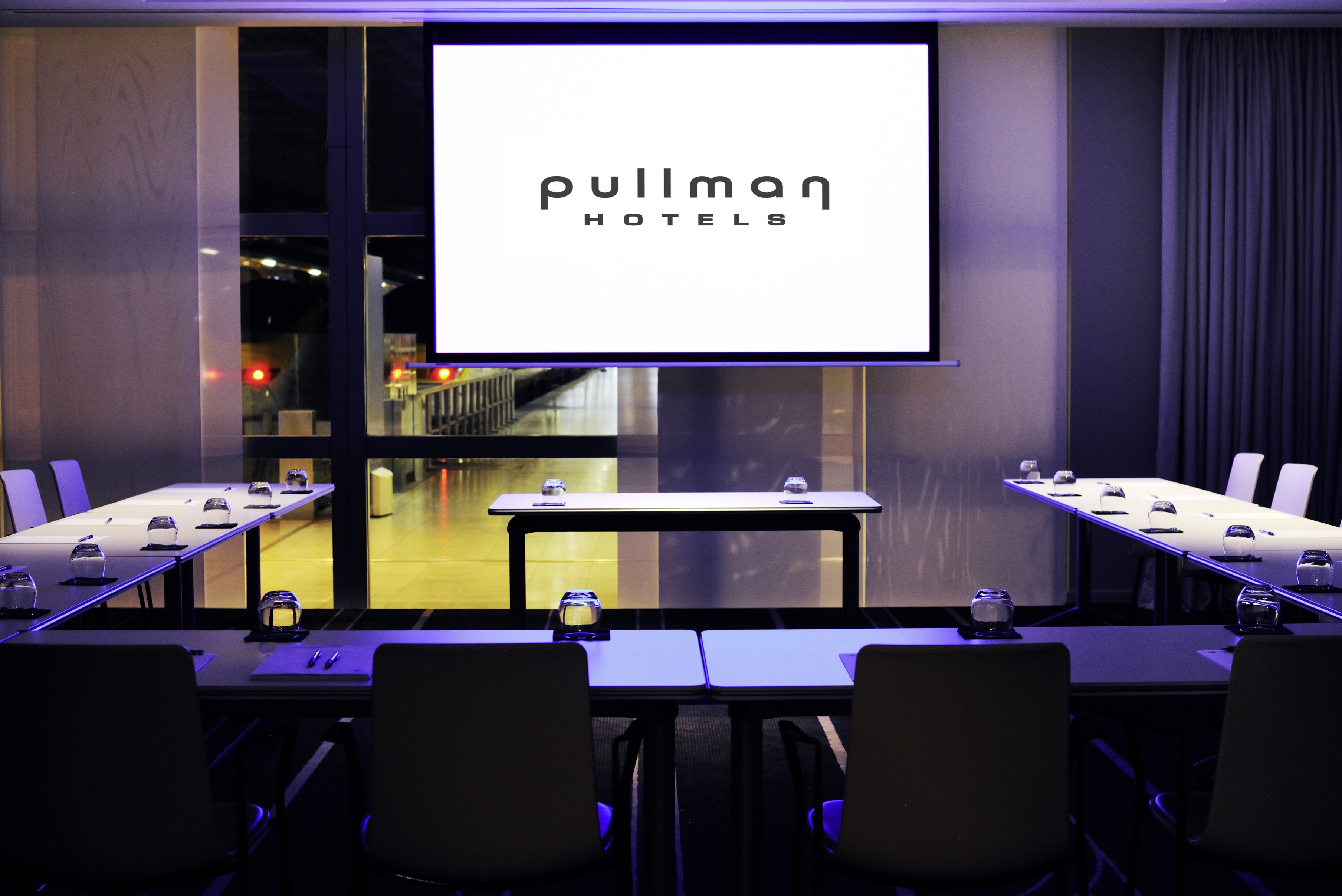 A conference room with a large screen displaying "Pullman Hotels" logo, a long table with chairs, and a view of the outside through large windows.