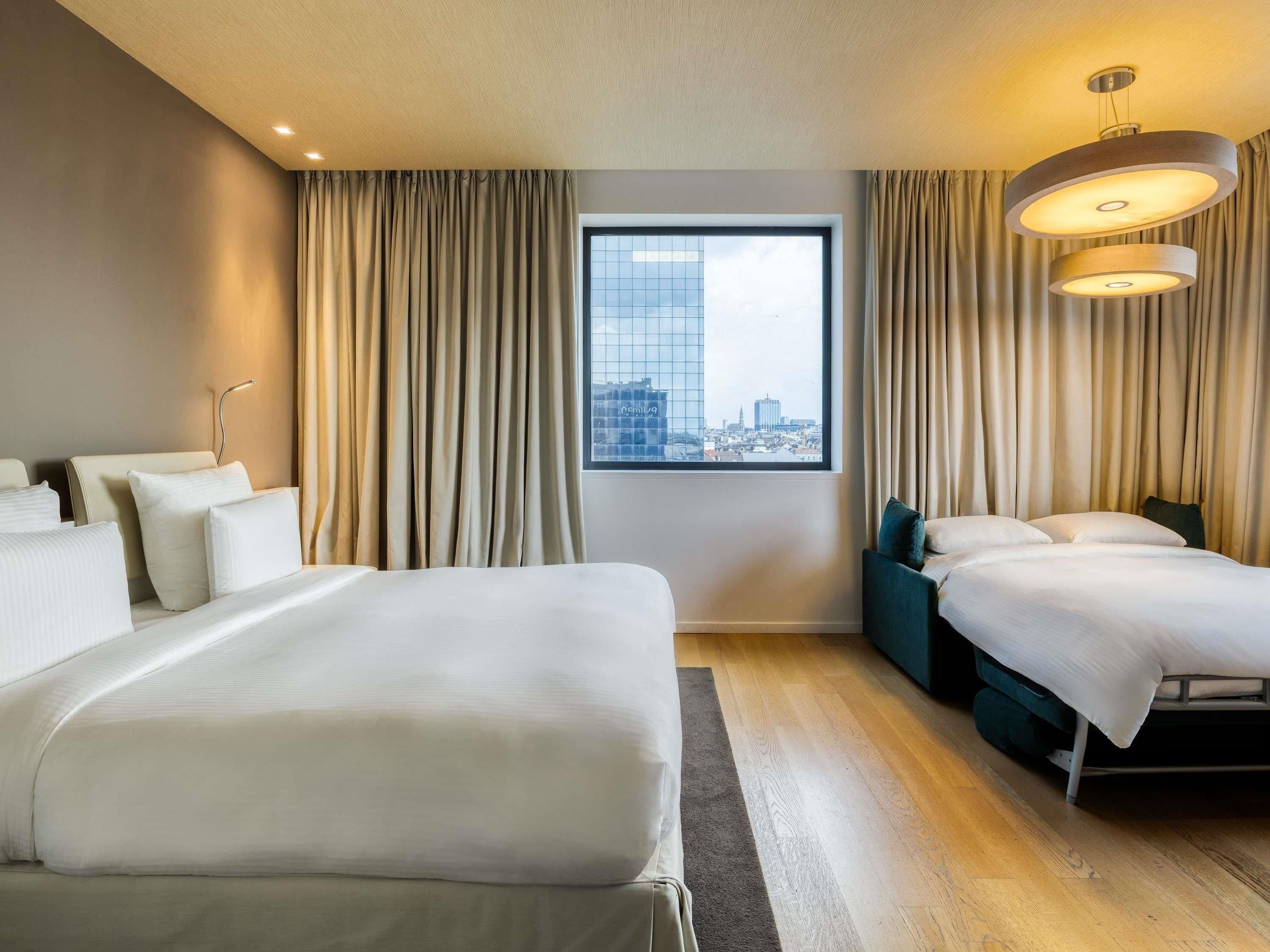 A hotel room with two beds, a large window with a city view, and a modern chandelier.