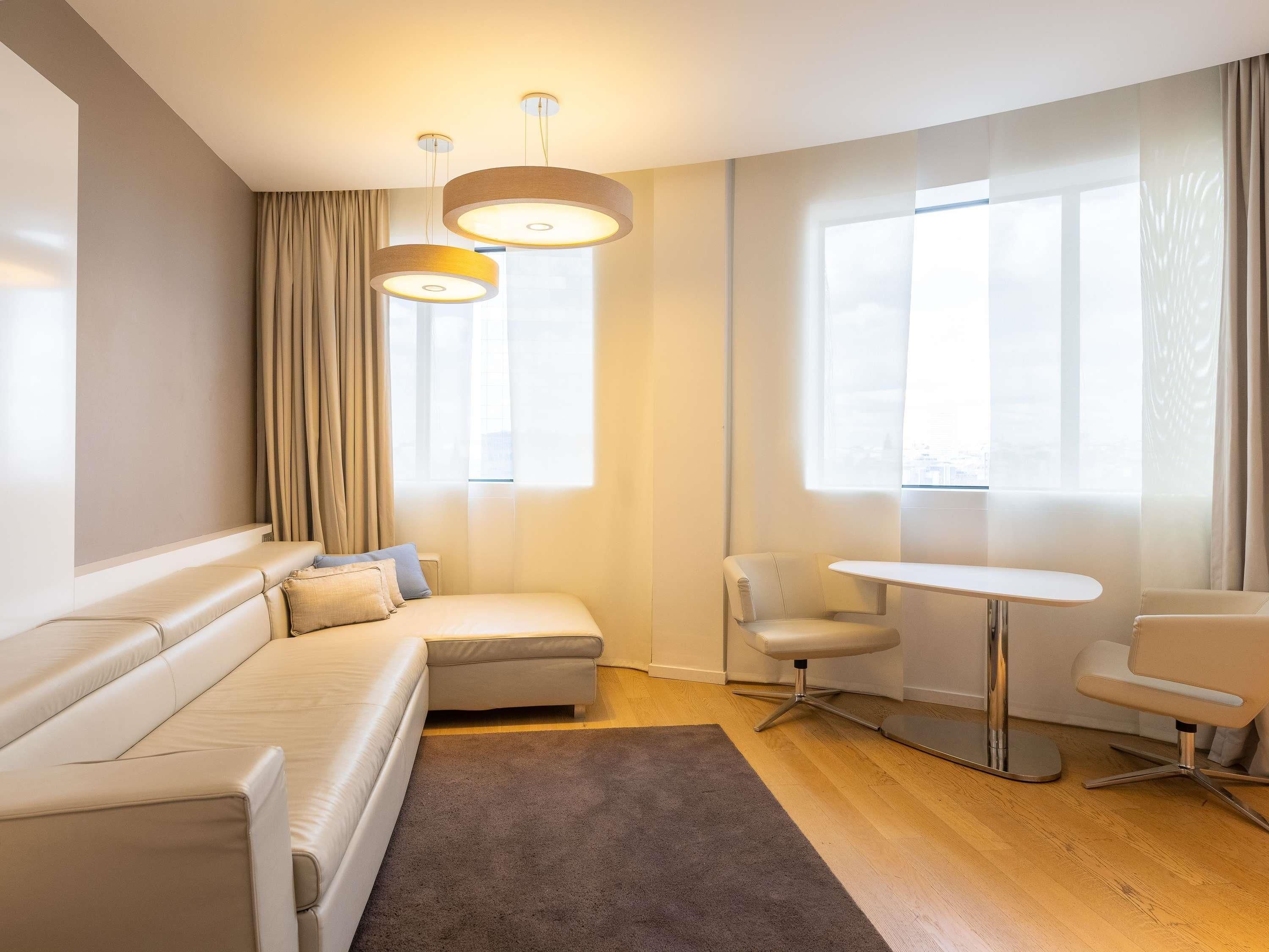 Una habitación de hotel moderna con sofá, una mesita, una silla y ventanas amplias.