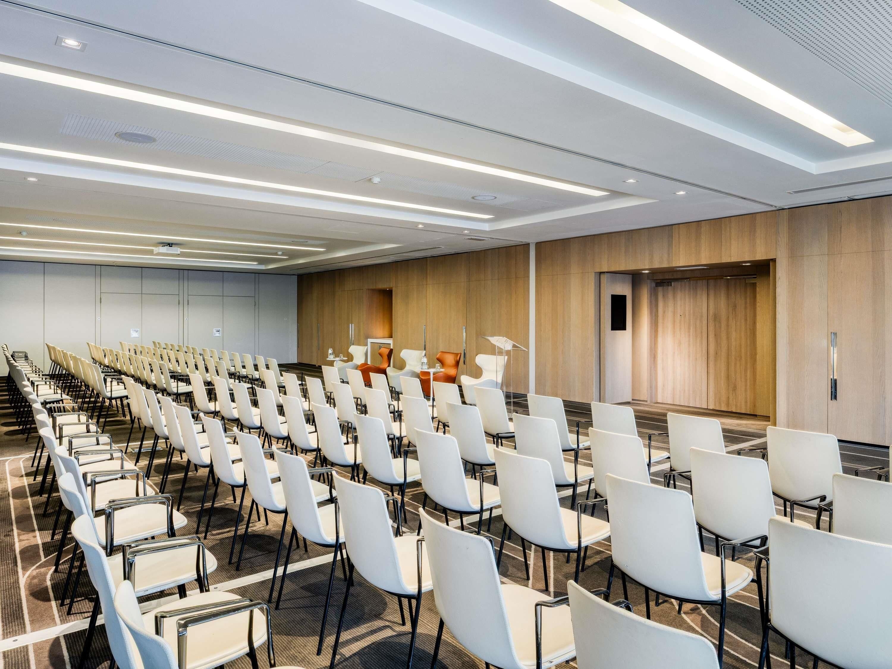 Un salón de conferencias con filas de sillas blancas, un panel de pared de madera e iluminación lineal en el techo.