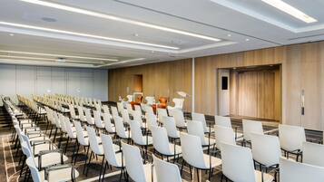 Un salón de conferencias con filas de sillas blancas, un panel de pared de madera e iluminación lineal en el techo.